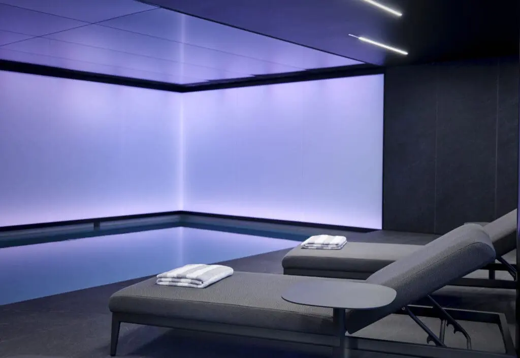 Indoor swimming pool at art'otel London Hoxton Old Street hotel with pool, featuring sleek gray loungers, folded towels, and soft purple ambient lighting.