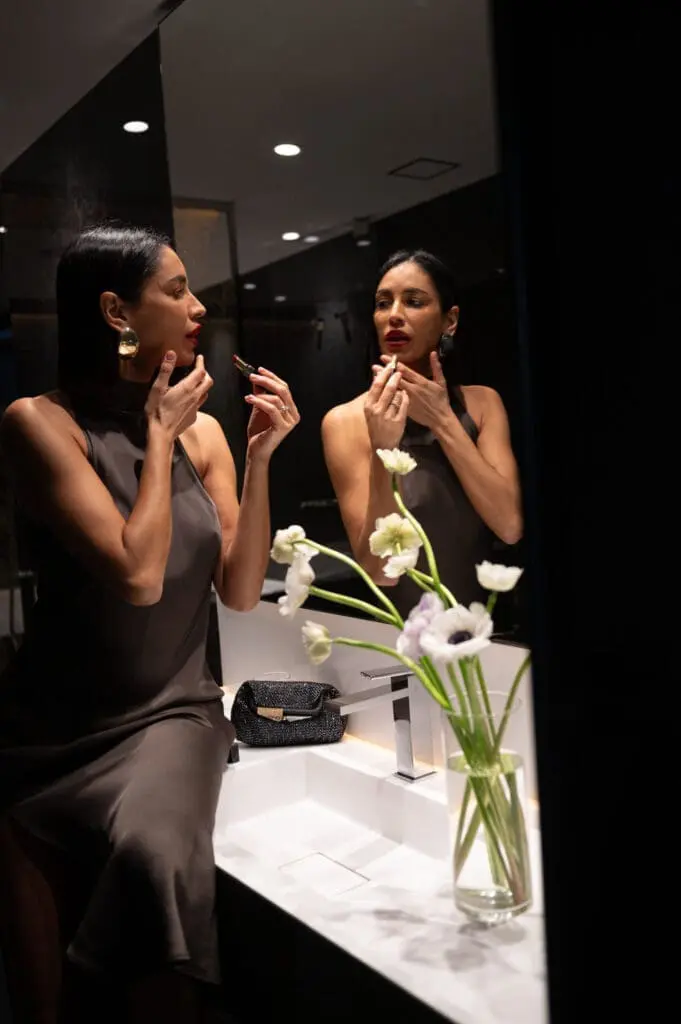Person applying lipstick in front of a mirror with white flowers and a black clutch on the counter at roma art rooms.
