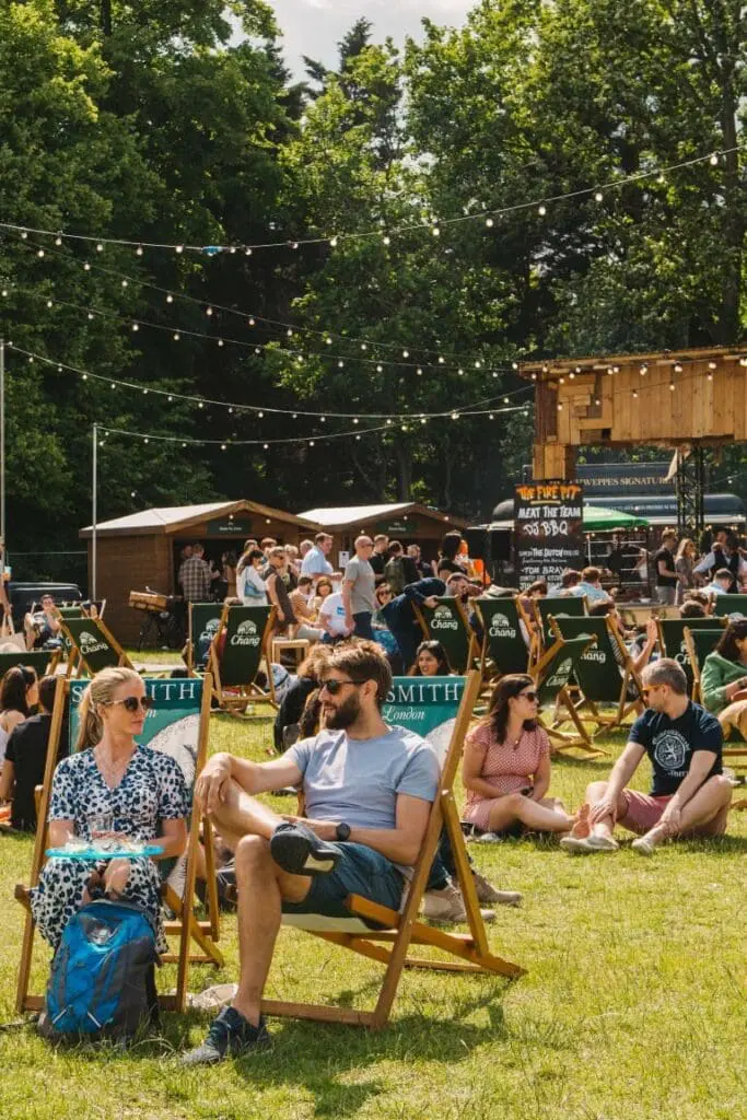 People relaxing on deck chairs at an outdoor summer festival surrounded by trees, food stalls and string lights in a lively park setting.
