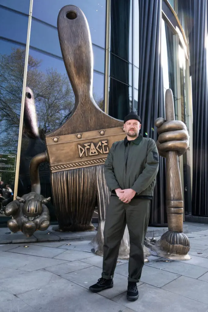 D*FACE standing in front his large bronze paintbrush sculpture outside art'otel London Hoxton Shoreditch hotel.