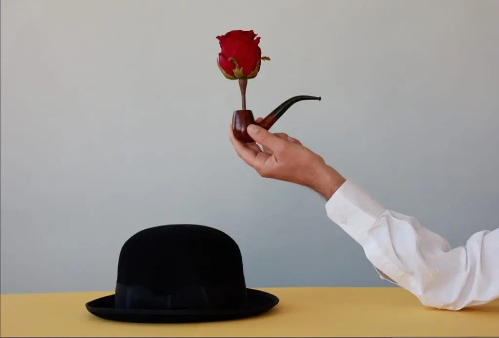 Person holding a pipe topped with a single red rose above a black bowler hat on a minimalist tabletop background.