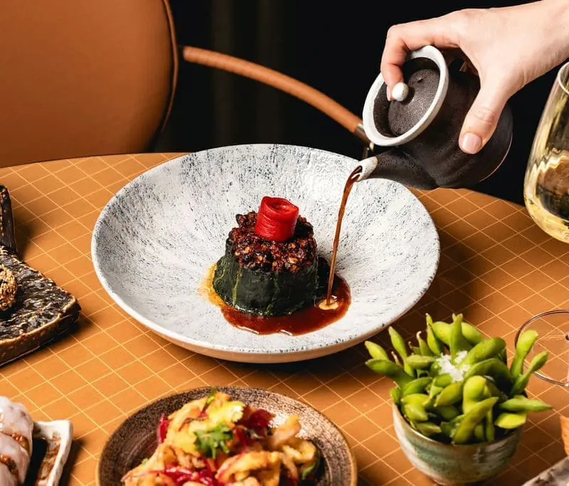 Hand pouring sauce from a black teapot onto a plated dish with vegetables, surrounded by edamame, wine glass, and other plates at art'otel Luxury art hotel Rome.