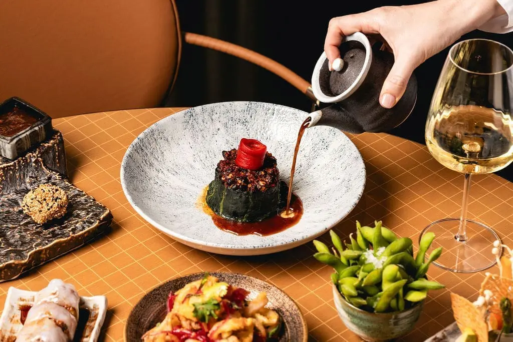 Elegant gourmet dish being served with sauce at art'otel Yezi restaurant Roma, surrounded by Asian delicacies and fine dining setup.