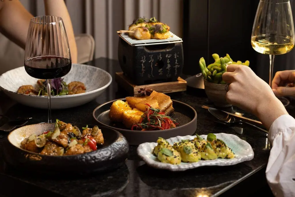 Elegant Japanese dining setup with assorted dishes, edamame, wine glasses, and modern table setting at art'otel Yezi restaurant Rome with gourmet dishes and wine.