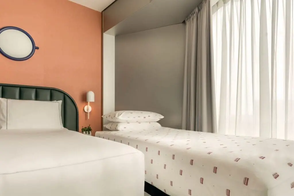 Contemporary twin bed setup at art'otel Battersea Power Station featuring stylish orange accent wall, modern lighting, and natural daylight.