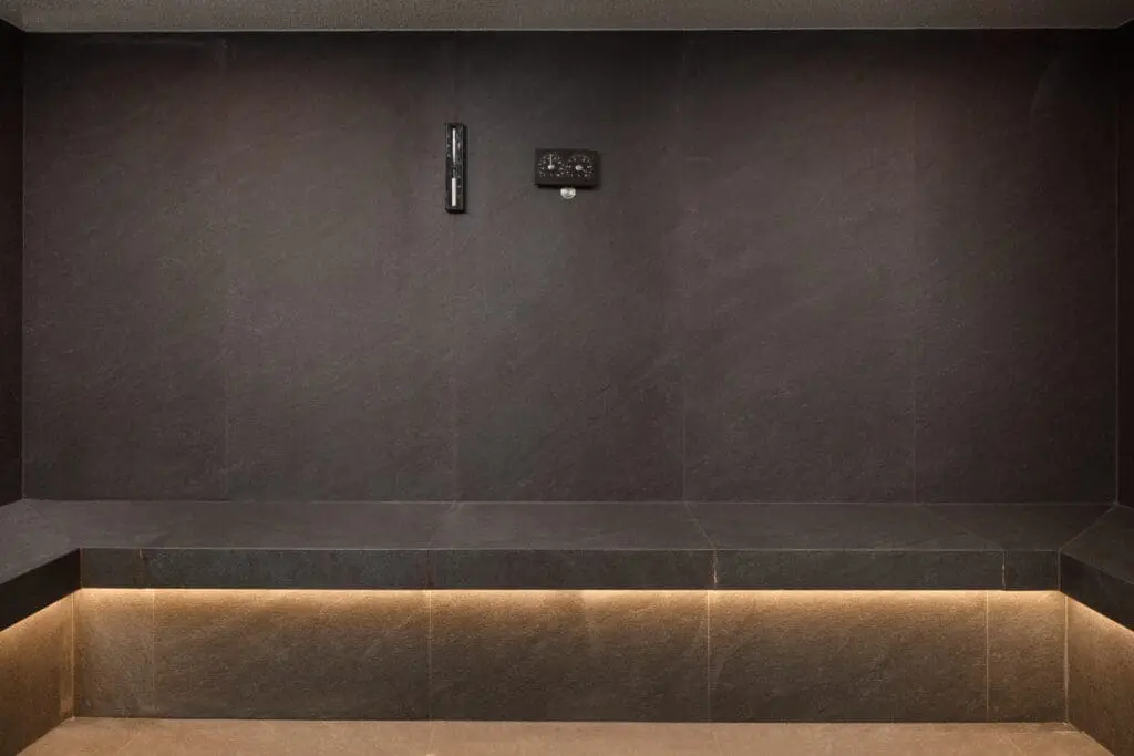Modern steam room with dark stone walls and built-in bench featuring warm underlighting at art'otel London Hoxton Shoreditch hotel with spa.