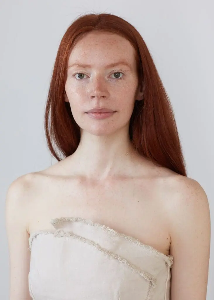 Person with long straight auburn hair wearing a strapless beige top with frayed edges, standing against a plain light gray background.