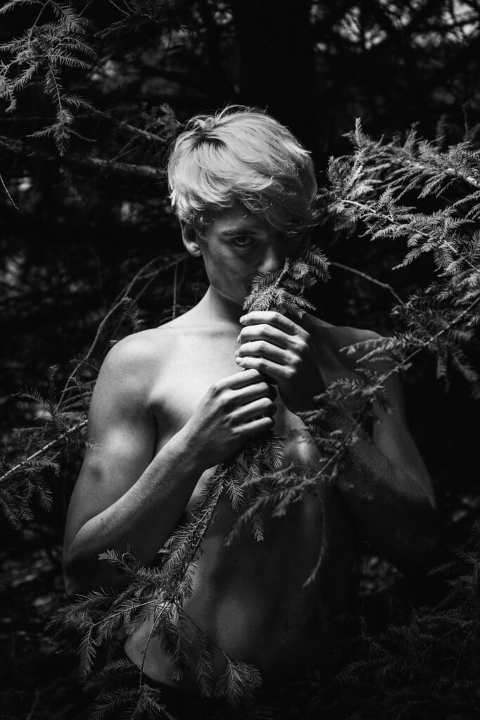 A naked blonde man hiding behind the branches of a tree.