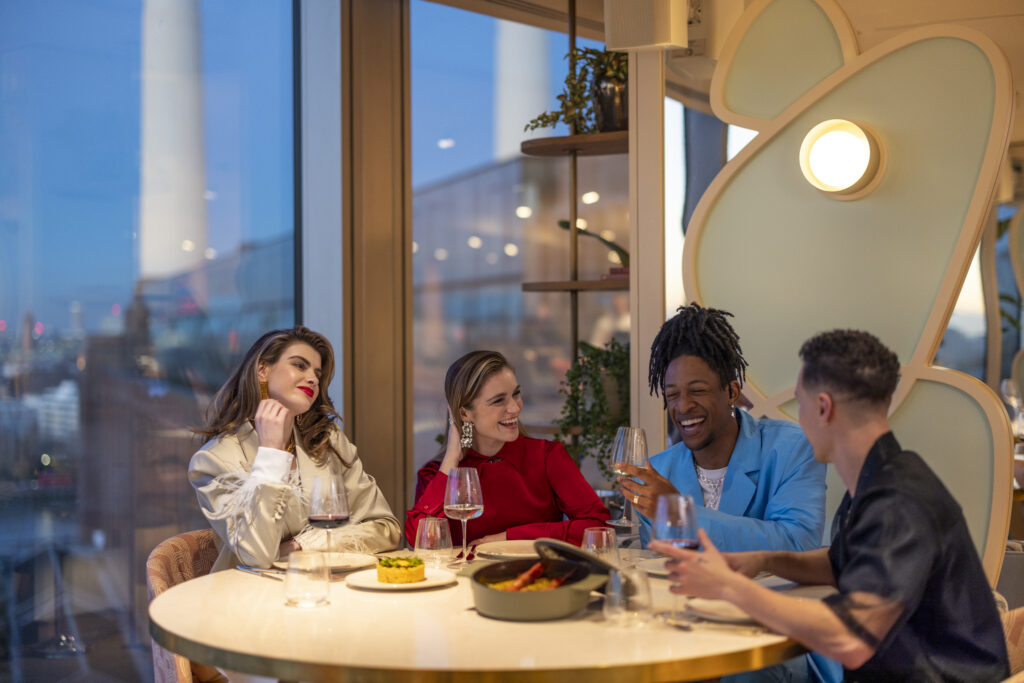 Four people sat at a table, all laughing and smiling, in JOIA with Battersea Power Station in the background.