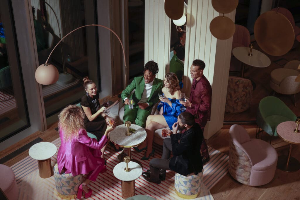 Group enjoying drinks in modern lounge with chic furniture, colorful outfits, stylish decor.