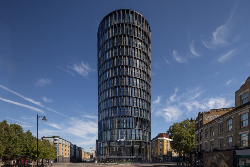 Art’otel London Hoxton Old Street – modern cylindrical glass hotel tower with spa, featuring reflective windows and contemporary architecture under a clear blue sky.